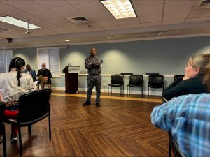 Man in black striped shirt with arms crossed standing on hardwood floor speaking to group of people with empty chairs in background