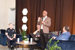 Man in brown coat speaking while standing holding a microphone as three men sit in chairs listening