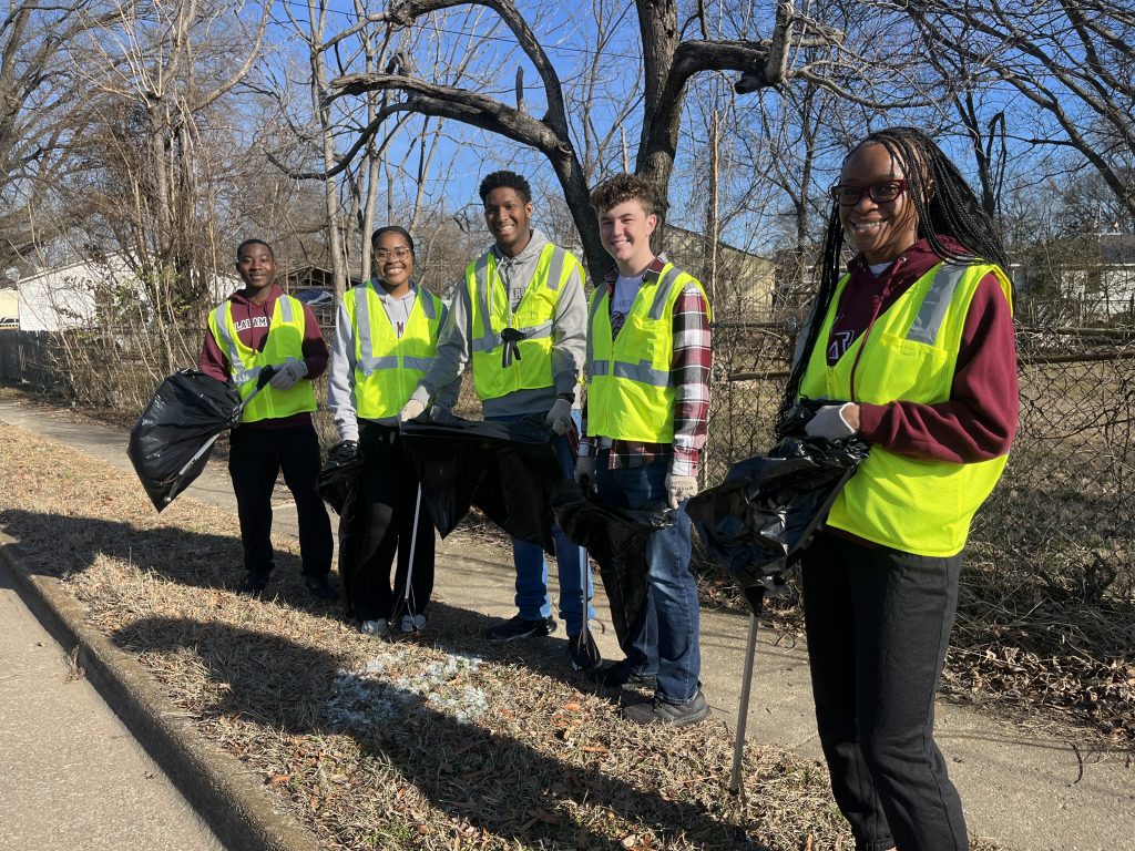 five people standingin a row outside wearing yellow vests and carrying garbage bags and litter pickup sticks
