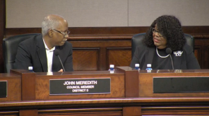 man sitting behind dais wearing glasses on left talks to woman wearing glasses on right in dark clothes