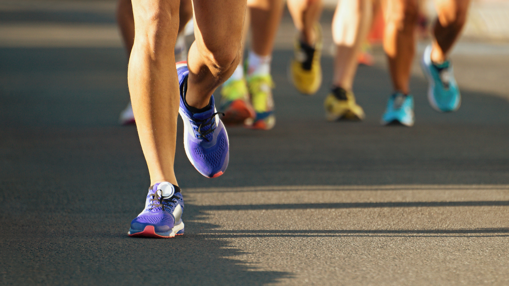 A group of runners in running shoes run down the street. Only their legs and shoes are visible.