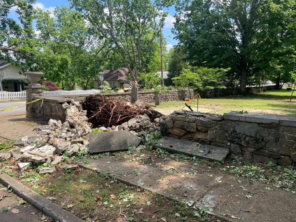 broken stone wall with debris on sidewalk with large green trees in background