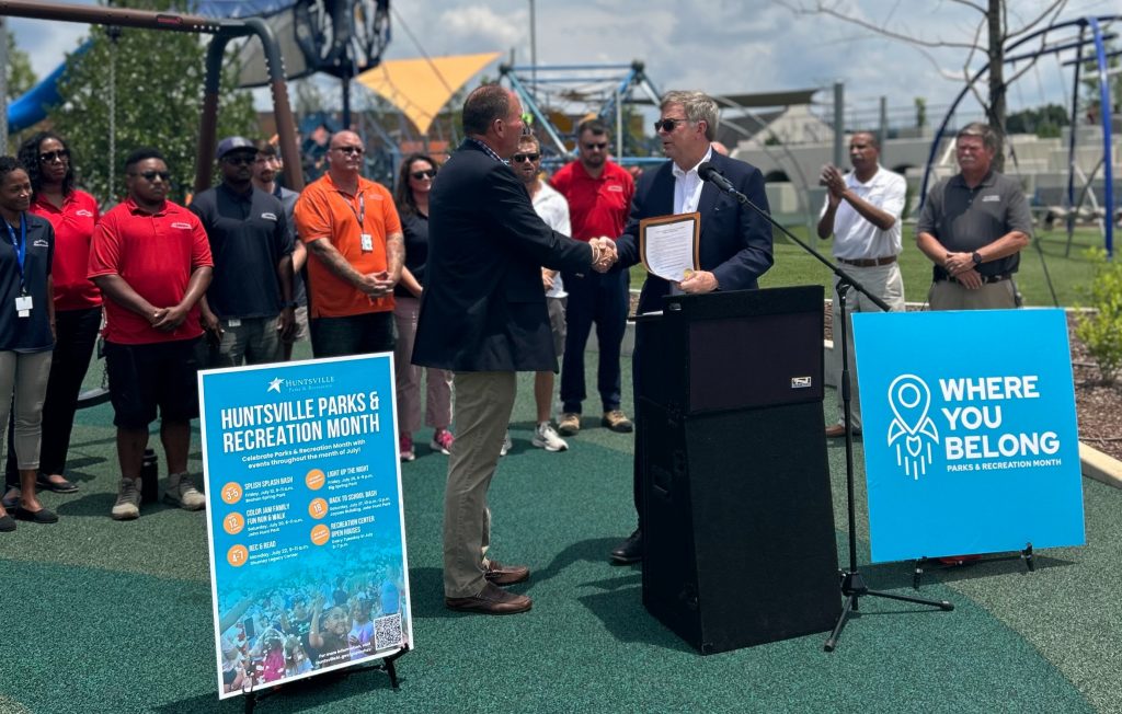 Mayor Tommy Battle, right, shakes hands with Huntsville Parks & Recreation Director James Gossett at a kickoff event for Parks and Recreation Month. The men are both wearing dark jackets. There are people standing to the rear in colorful shirts. There is a playground in the background.
