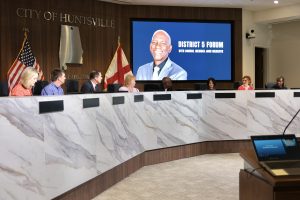 group of people seated behind dais with Alabama flag and video screen in background