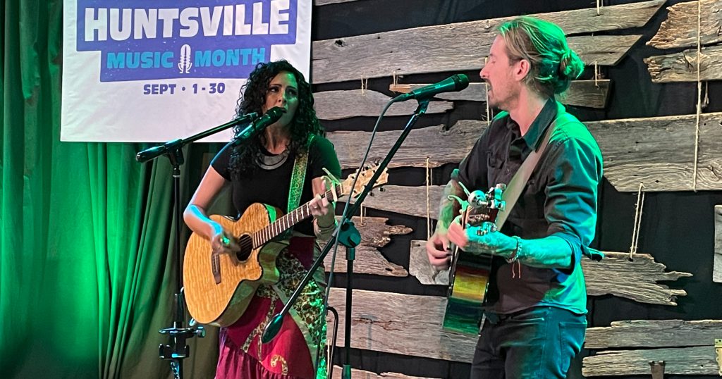 Huntsville duo Luna Koi perform at the 2024 Huntsville Music Month kickoff at Tangled String Studios. The woman at left is wearing a dress and the man at right a dark shirt and dark jeans. They are both playing acoustic guitars.