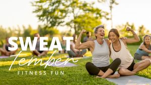 Two women sitting on an exercise mat hug. They are both flexing their arms while laughing. There are trees in the background.