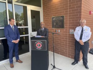 man in dark suit speaking at podium with man in suit on left and man in uniform with white shirt on right standing in front of brick wall