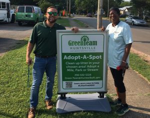 man and woman standing on sidewalk next to green and white adopt-a-spot sign