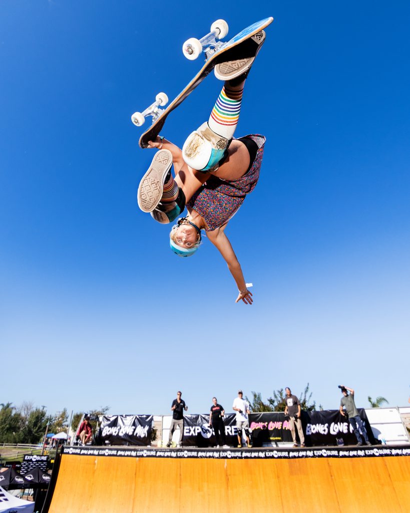 A female skater is in the air on a skateboard while people in the background watch.