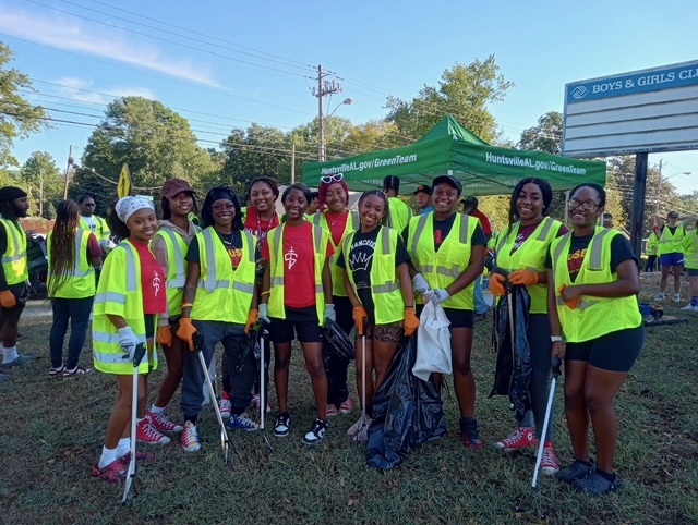 Group of women outdoors wearing green safety vests with blue sky and trees in background