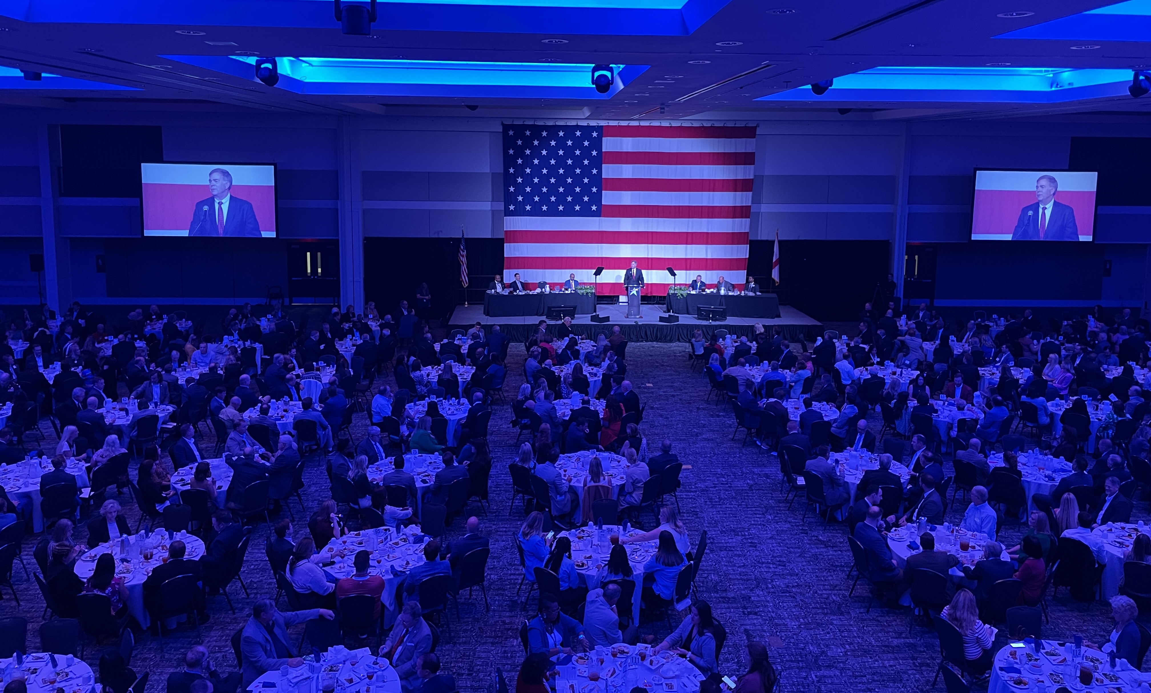 large ballroom with people at tables with American flag in background