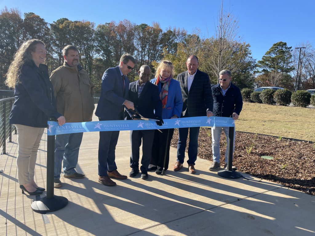 group of people wearing winter coats lined behind ribbon for ribbon cutting while standing on sidewalk