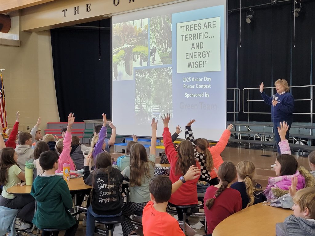 children raising hands at school assembly looking at projection screen with display about trees