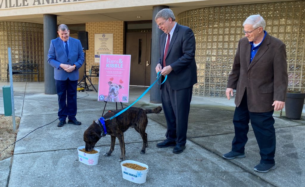 Three men in suits watch a dog eat from a bowl in front of Huntsville Animal Services. There is a pink poster that says Kisses and Kibble in the background.
