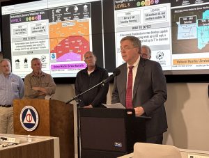 man in coat and tie speaks at podium with digital display screens in background