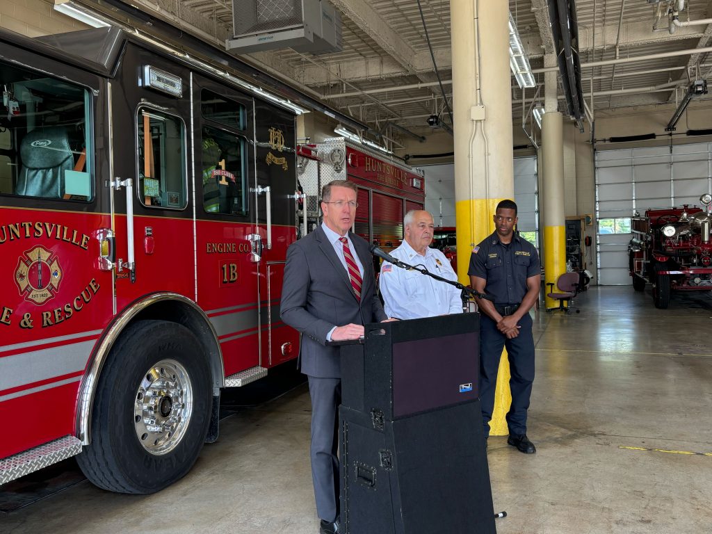 man in suit speaking at podium with fire truck in background