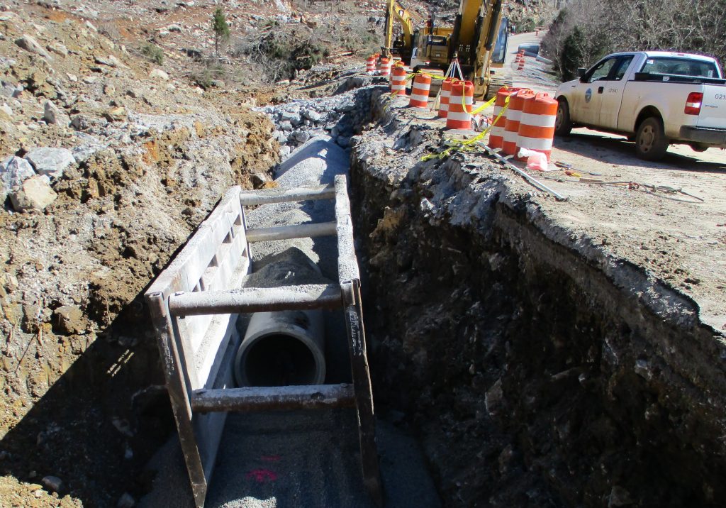 construction site with large pipe laying in ditch with orange traffic barrels and white pickup truck in background