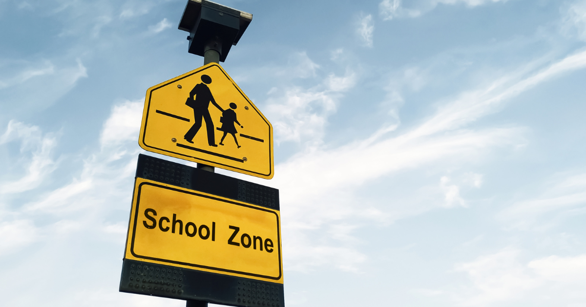 A yellow road sign shows the silhouette of an adult holding the hand of a child as they walk across a crosswalk. Beneath it, a second yellow sign reads “School Zone.” Both signs are mounted on a pole against a bright sky with wispy clouds.