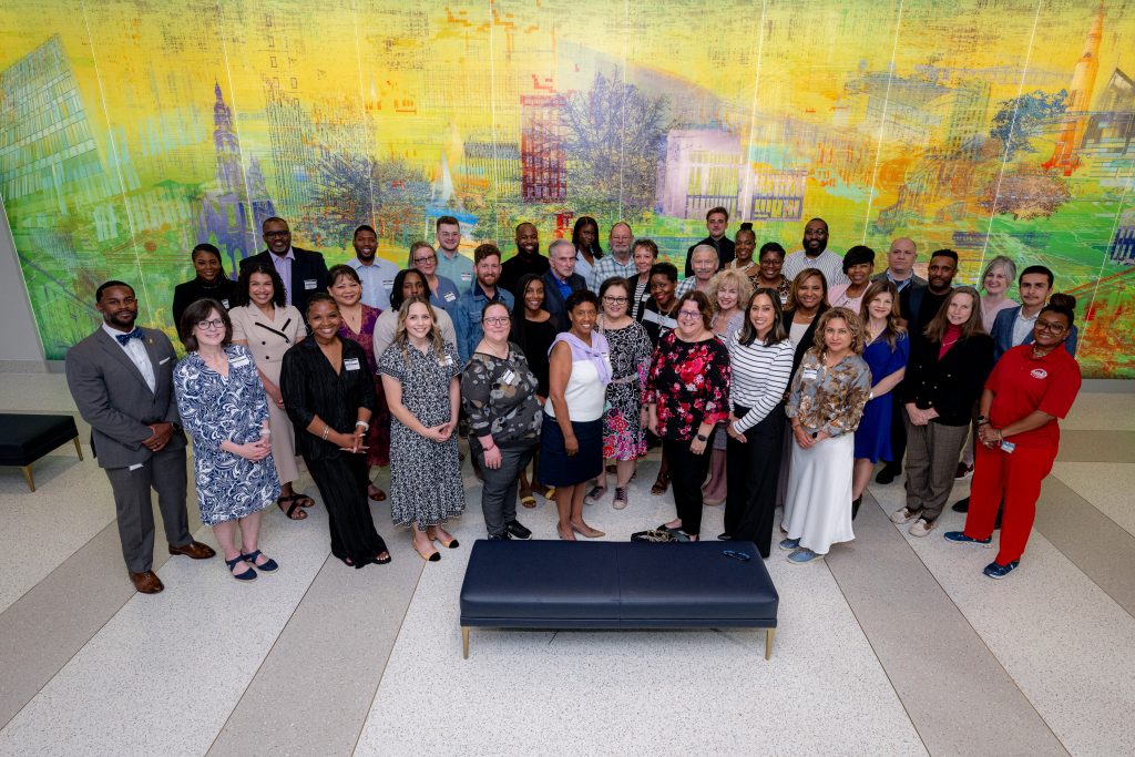Group photo of 50 graduates from the Civic Engagement Academy spring 2025 cohort in front of digital artwork in Huntsville City Hall