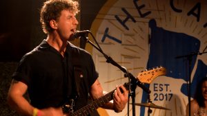 Photo of a white male guitar player singing into a mic onstage at The Camp at MidCity