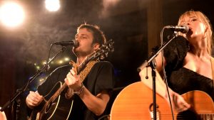 Photo of a white male guitar player and a white female guitar player singing into a mic onstage at The Camp at MidCity