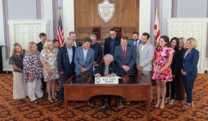 Group of 17 men and women looking down at Alabama Governor Kay Ivey at a ceremonial bill signing in the Alabama State Capitol