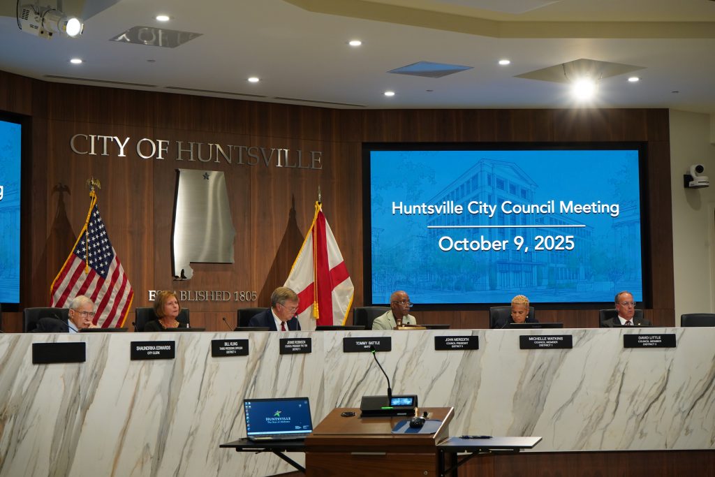city council sitting at white dais with flags and projection screens in background
