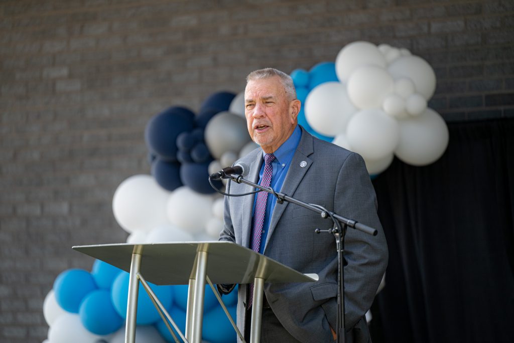 man in a suite and tie stands at a podium in front of a blu and white balloon arch