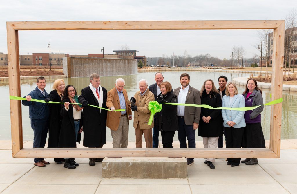 group of people standing outdoors in a picture frame in front of a lake, holding a green ribbon for a ribbon-cutting ceremony