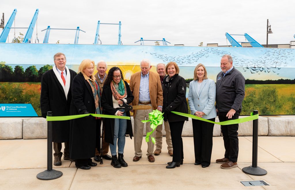 Group of people standing in line in front of outdoor fitness equipment, holding a green ribbon for a ribbon cutting