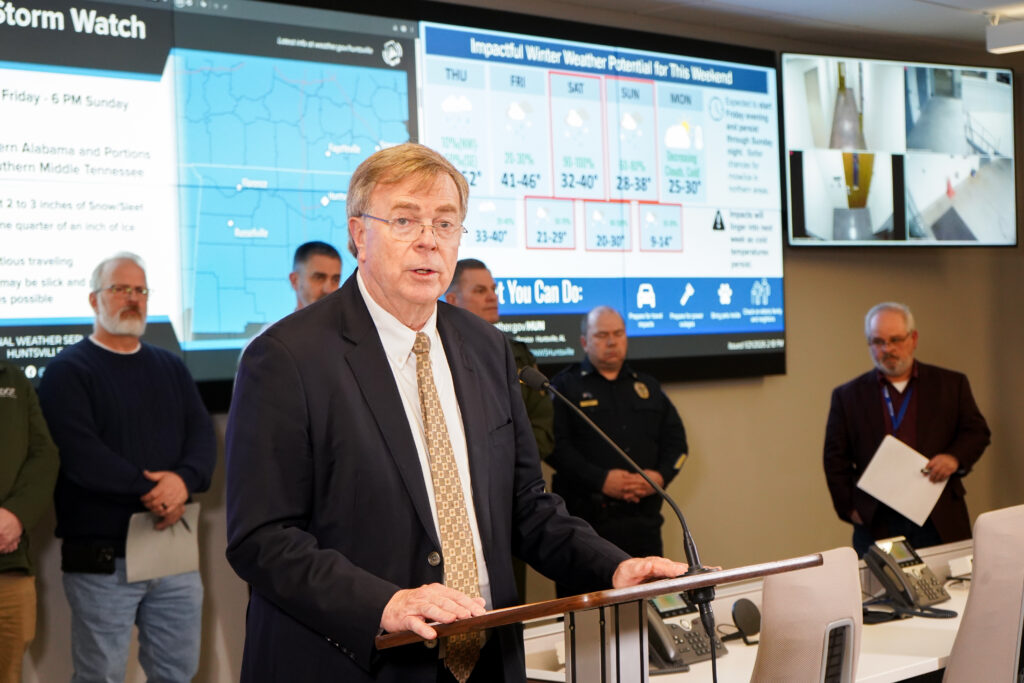 man in dark suit speaks at podium with projection screens in background