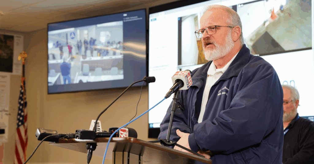 White male standing, talking at podium with microphones from media in front of monitors
