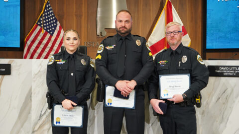 Three officers smiling at the camera in this photo, holding their awards they were presented at a City Council meeting