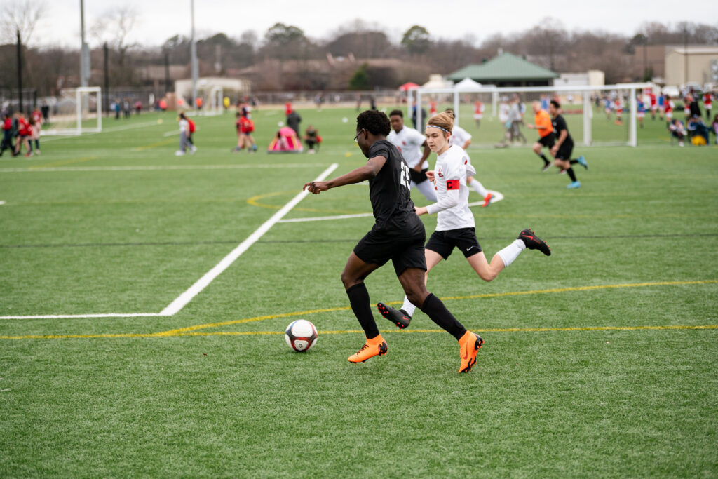 Two young mean racing to kick a rolling soccer ball with a crowd of players and fans in the background.