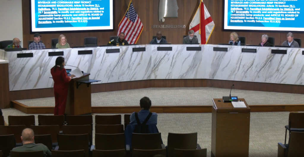 group of people sit at a large marble desk facing an audience. Two large TV screens are behind them.