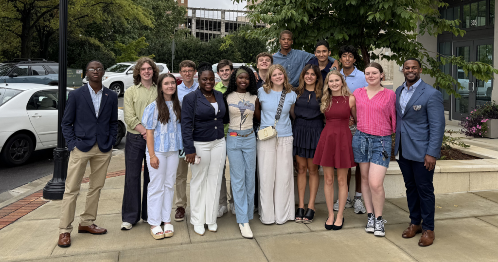 A group of young people and a man dressed in a suit stand together outside Huntsville City Hall, with cars and trees in the background.