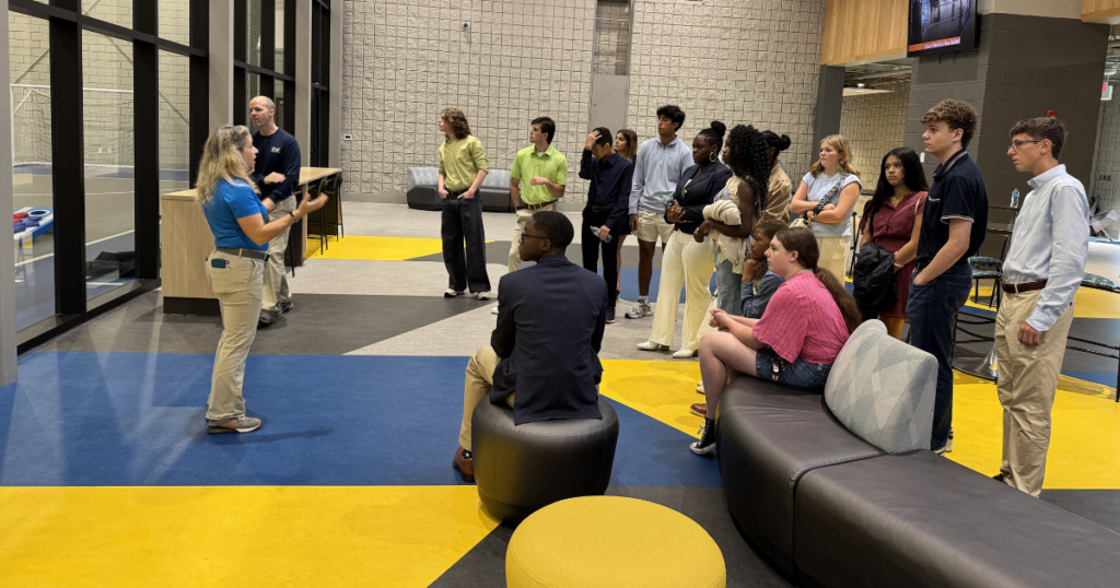 A woman in a blue shirt and khaki pants gestures while speaking to a group of high school aged students standing and sitting in a modern room with blue and yellow flooring.