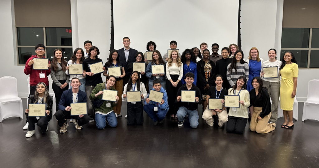 A group of high school aged students are posing for a photo holding certificates. They are standing and kneeling in rows, with a projector screen in the background.