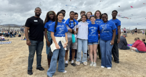 A group of high school aged students and a man pose for a photo at a community kite festival, with kites visible in the cloudy sky and other festival-goers in the background.