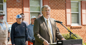 A man in a suit and tie speaks into a microphone in front of a brick building, with several other men standing behind him.