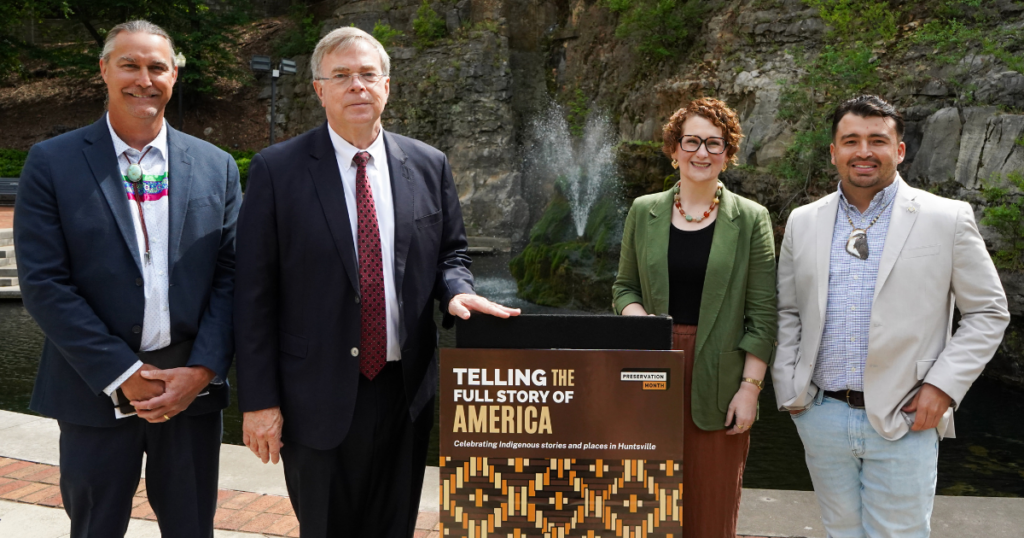 Four individuals stand in front of a spring and rocky cliff. In the foreground, a display reads "TELLING THE FULL STORY OF AMERICA" and "Celebrating Indigenous stories and places in Huntsville." The individuals are posing for a photograph.