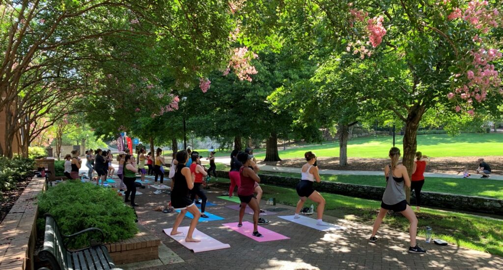 A group of adults participates in an outdoor fitness class in a shaded park, standing on yoga mats and following an instructor while exercising. Trees with green leaves and pink blossoms surround the paved area, and sunlight filters through the canopy onto the participants.