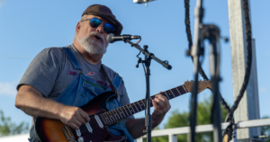 A man wearing overalls, a cap, and sunglasses plays a sunburst electric guitar while singing into a microphone on an outdoor stage.