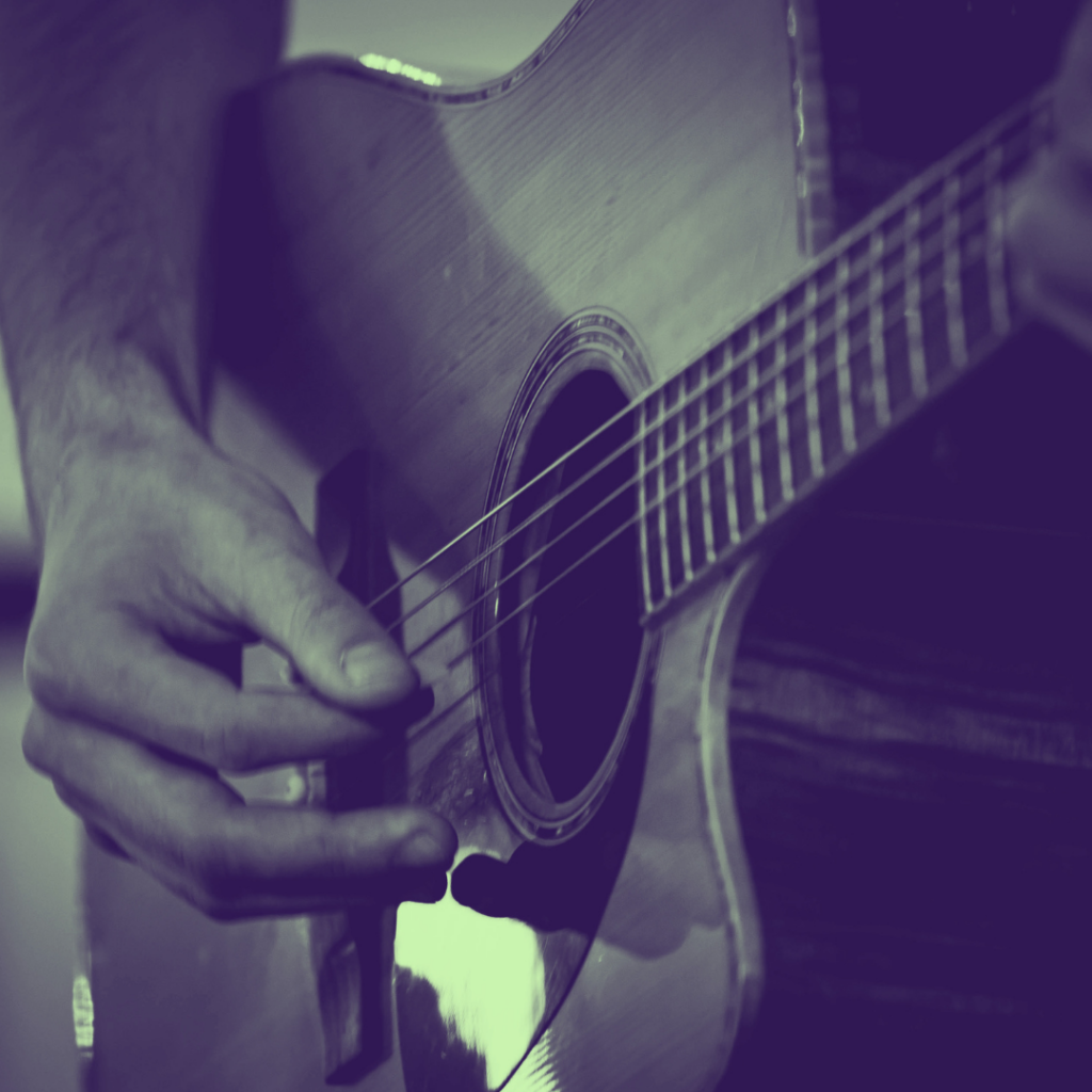 Close‑up of a person playing an acoustic guitar, with their hand strumming the strings near the sound hole. The wooden body of the guitar is visible, and the lighting creates a warm, moody atmosphere.