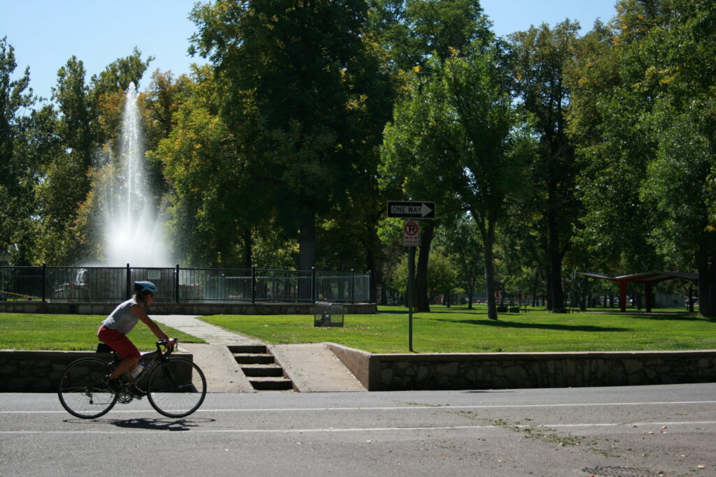 A cyclist rides their bike on the road. The rider is wearing a helmet and there is a fountain in the background.