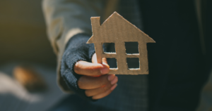 A hand holds a small, cutout cardboard house with square windows and a peaked roof.