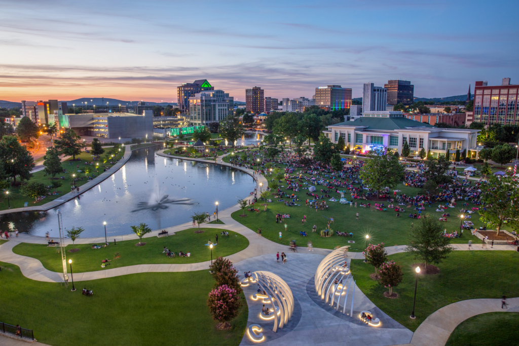 Aerial view of a large city park at sunset, showing a pond with a fountain, winding pathways, and expansive lawns filled with groups of people gathered for an outdoor event. City buildings surround the park, illuminated by evening light, with colorful lighting features and a performance area visible near the center.