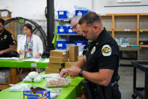 A uniformed law enforcement officer stands at a worktable, inspecting a clear plastic bag filled with rubber bands. Several cardboard packages, storage bags and other supplies are spread across the bright green table. In the background, additional individuals work at the same table, and shelves stocked with blue storage bins and various items line the wall of the warehouse-like workspace.