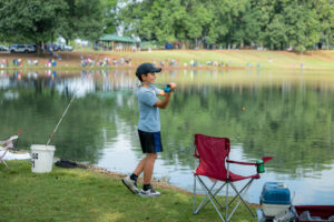 A child smiles and casts a fishing pole into a pond. It appears to be a sunny day and there are other groups of people in the background also fishing.