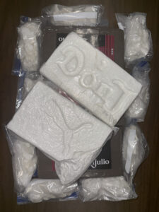 A group of tightly wrapped plastic packages containing white, powdery or rock‑like substances arranged on a wooden surface. Two large rectangular white blocks with textured surfaces lie on top, one showing an embossed decorative pattern and the other showing an embossed flowing line. Several smaller sealed plastic bags filled with similar white material surround the larger blocks. A cardboard box labeled “Don Julio” is partially visible beneath the items.